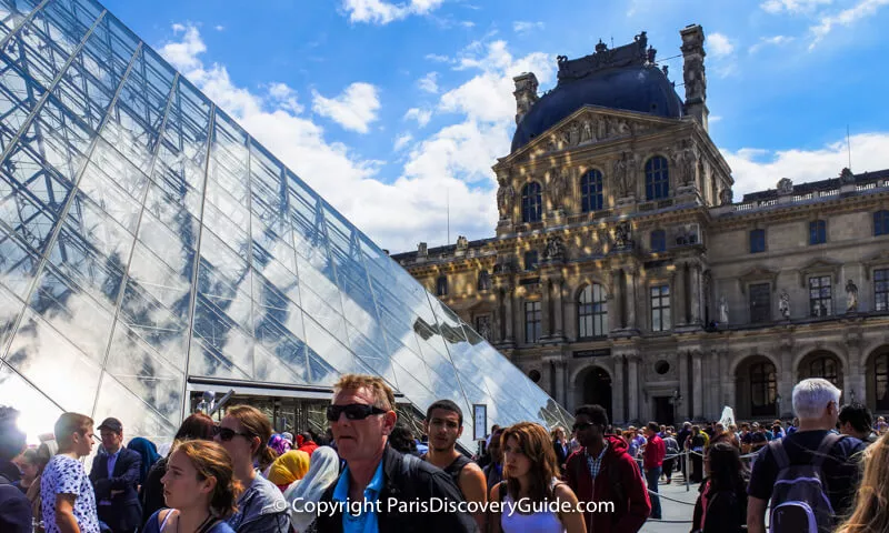 Ticket line at the Louvre winding around the courtyard; nearby signs warn of a 2+ hour wait